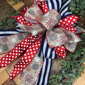 May include: A patriotic bow featuring red and white polka dot ribbon, navy blue and white striped ribbon, and ribbon with a firework design. The bow is set against a wooden background with greenery.