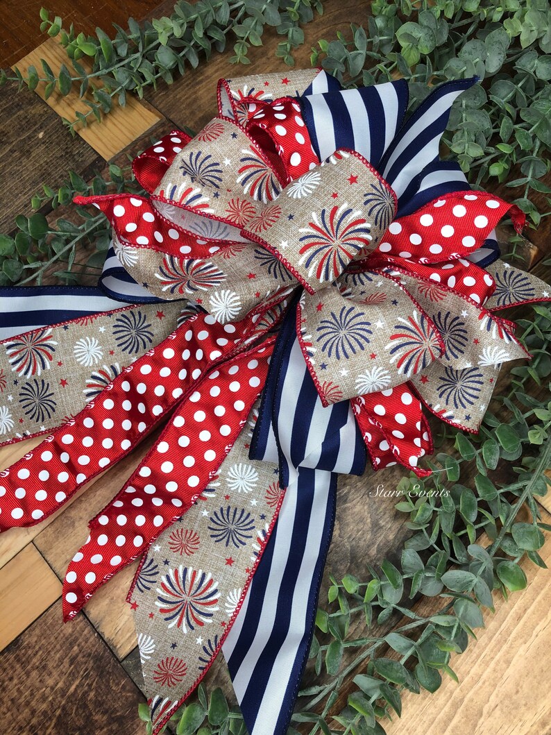 May include: A patriotic bow featuring red and white polka dot ribbon, navy blue and white striped ribbon, and ribbon with a firework design. The bow is set against a wooden background with greenery.