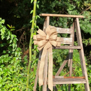 May include: A large, brown burlap bow with long tails is tied to a wooden ladder. The bow is tied in a knot with a loop at the top. The ladder is made of wood and has three steps. The bow is sitting on the top step of the ladder. The background is a green bush.