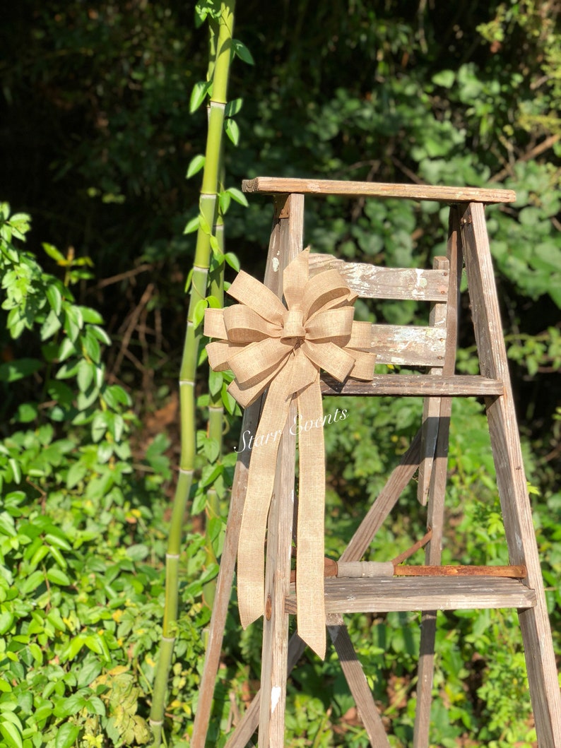 May include: A large, brown burlap bow with long tails is tied to a wooden ladder. The bow is tied in a knot with a loop at the top. The ladder is made of wood and has three steps. The bow is sitting on the top step of the ladder. The background is a green bush.