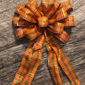May include: A large orange and green plaid bow with gold accents. The bow has four tails and is tied in a knot. The bow is on a wooden surface.
