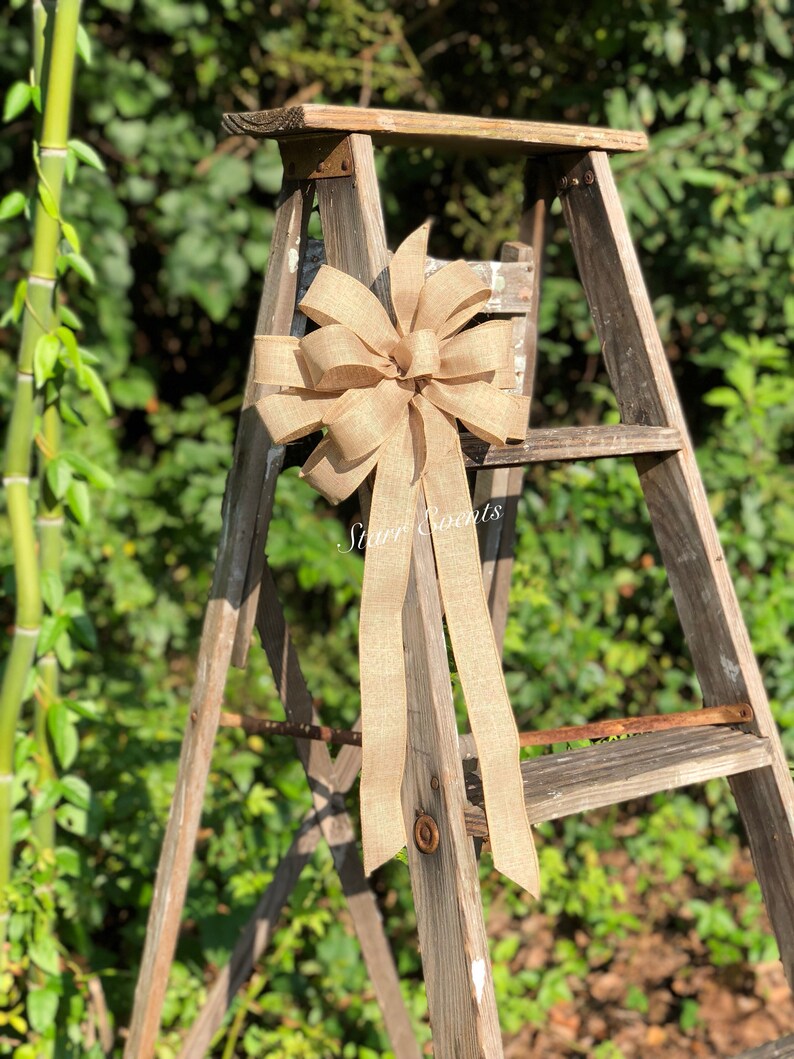 May include: A large, rustic, brown burlap bow with long tails is tied to a wooden ladder. The bow is tied to the top rung of the ladder, and the tails hang down the side of the ladder.