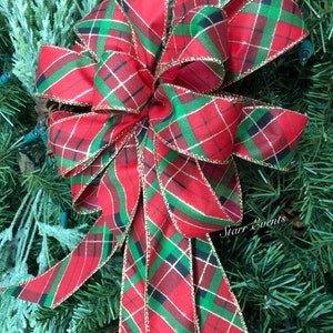 May include: A large, decorative bow with a red, green, and black plaid pattern. The bow has gold trim and is set against a backdrop of green pine needles. The bow is likely for holiday or seasonal decor.