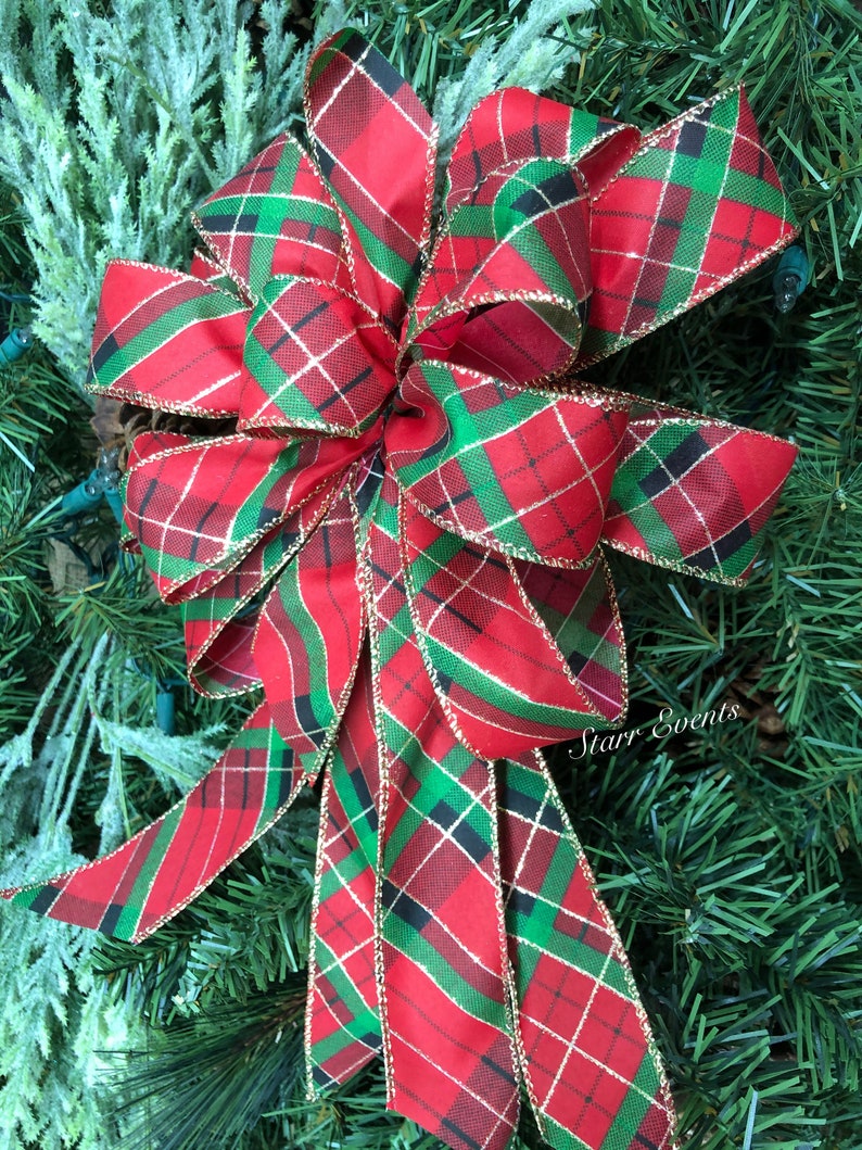 May include: A large, decorative bow with a red, green, and black plaid pattern. The bow has gold trim and is set against a backdrop of green pine needles. The bow is likely for holiday or seasonal decor.