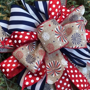 May include: A patriotic bow featuring red and white polka dot ribbon, navy blue and white striped ribbon, and ribbon with a firework design. The bow is set against a backdrop of green foliage.