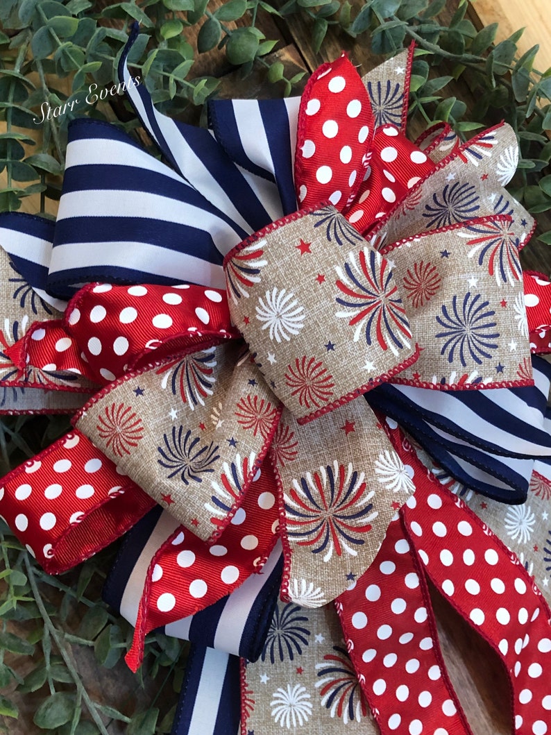 May include: A patriotic bow featuring red and white polka dot ribbon, navy blue and white striped ribbon, and ribbon with a firework design. The bow is set against a backdrop of green foliage.