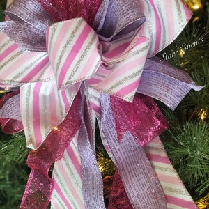 K&ouml;nnte beinhalten: Ein rosa-wei&szlig; gestreiftes Band mit silbernem Glitzer, ein rosa glitzerndes Band und ein lavendelfarbenes glitzerndes Band sind zu einer Schleife gebunden. Die Schleife befindet sich auf einem gr&uuml;nen Weihnachtsbaum.