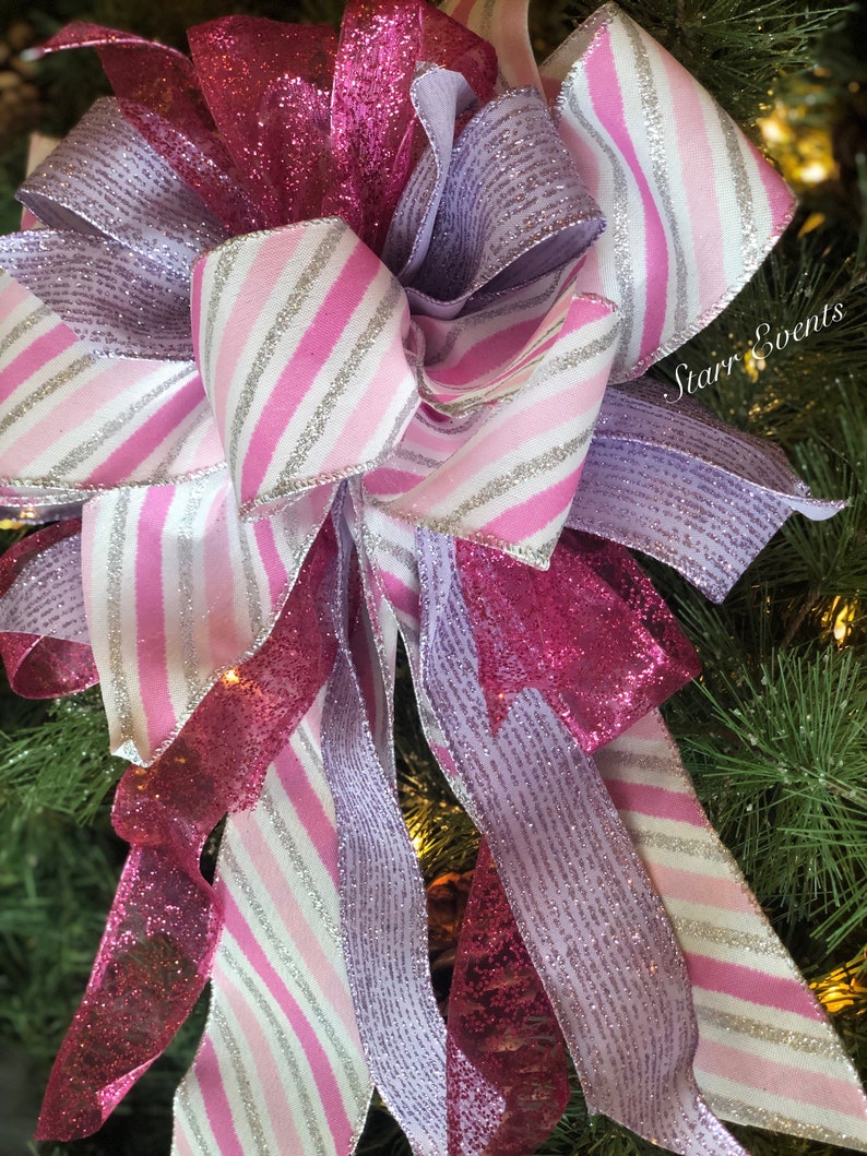 K&ouml;nnte beinhalten: Ein rosa-wei&szlig; gestreiftes Band mit silbernem Glitzer, ein rosa glitzerndes Band und ein lavendelfarbenes glitzerndes Band sind zu einer Schleife gebunden. Die Schleife befindet sich auf einem gr&uuml;nen Weihnachtsbaum.