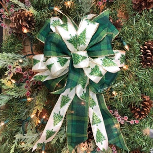 May include: A festive Christmas bow with a green plaid pattern and white ribbon featuring green Christmas trees. The bow is surrounded by pine needles, pine cones, and red berries, creating a holiday theme.