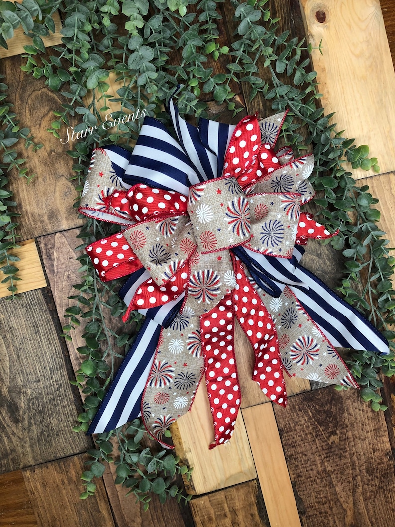May include: A patriotic bow featuring red and white polka dots, navy blue stripes, and a beige ribbon with firework designs. The bow is set against a wooden background with greenery, perfect for festive decorations. The text "Starr Events" is visible.