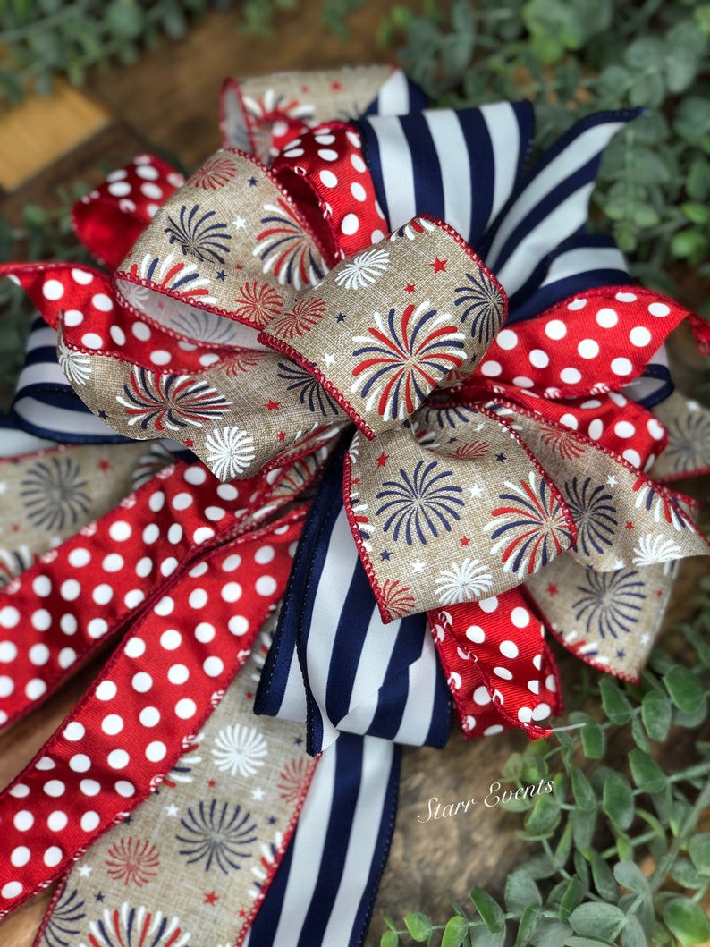 May include: A decorative bow featuring patriotic colors and patterns. The bow includes red and white polka dot ribbon, navy blue and white striped ribbon, and tan ribbon with a firework design. The bow is suitable for various decorative purposes.