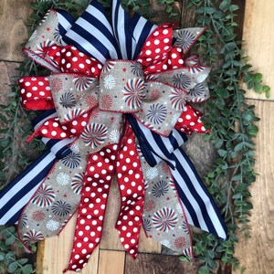 May include: A patriotic bow featuring red, white, and blue ribbons. The bow has a mix of patterns, including red polka dots, navy blue stripes, and a tan ribbon with firework designs. The text "Starr Events" is visible.
