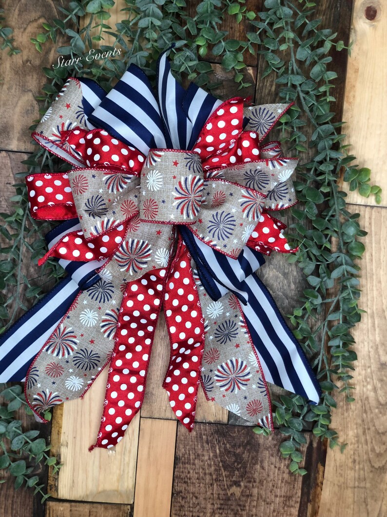 May include: A patriotic bow featuring red, white, and blue ribbons. The bow has a mix of patterns, including red polka dots, navy blue stripes, and a tan ribbon with firework designs. The text "Starr Events" is visible.