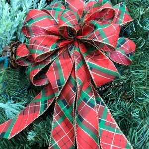 May include: A large, decorative Christmas bow with a red, green, and gold plaid pattern. The bow is made of fabric and has long, flowing tails. The bow is set against a backdrop of green pine needles and a pine cone.