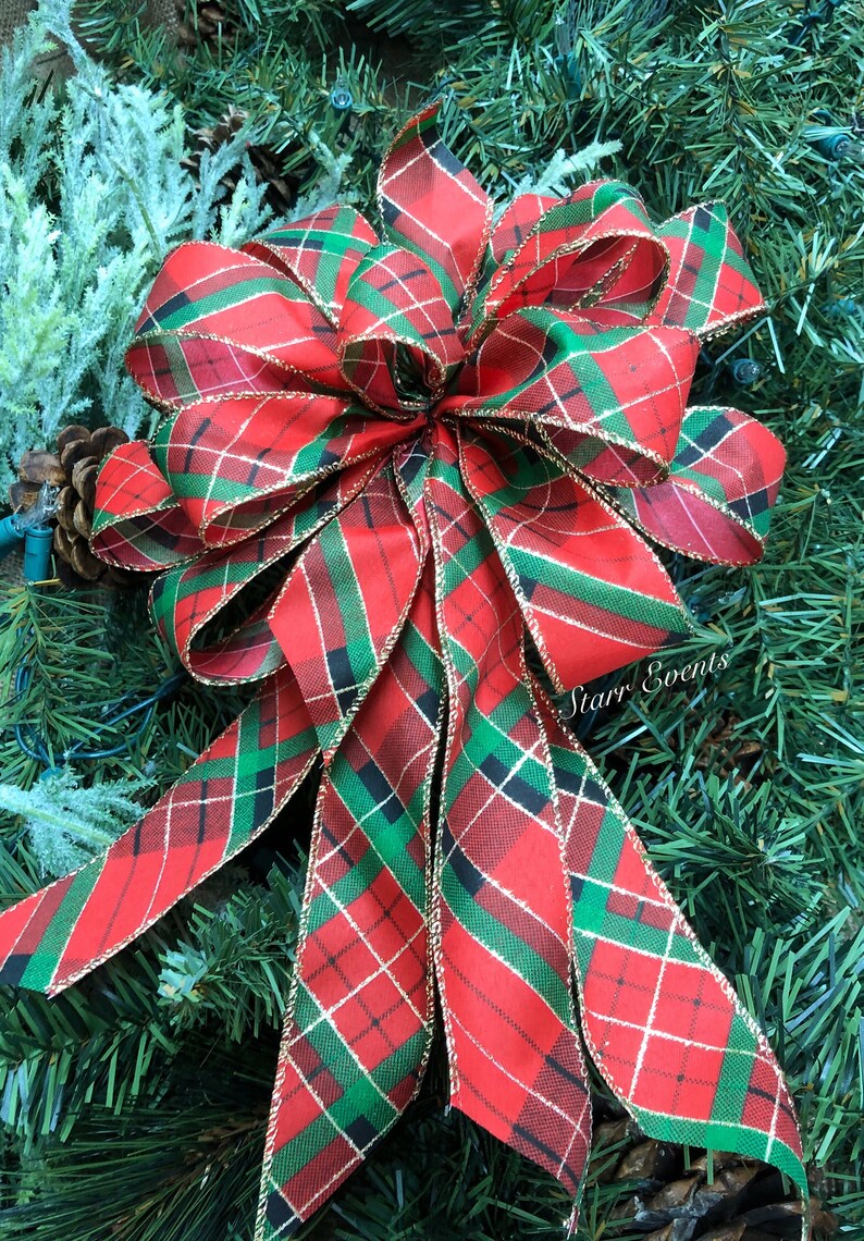 May include: A large, decorative Christmas bow with a red, green, and gold plaid pattern. The bow is made of fabric and has long, flowing tails. The bow is set against a backdrop of green pine needles and a pine cone.