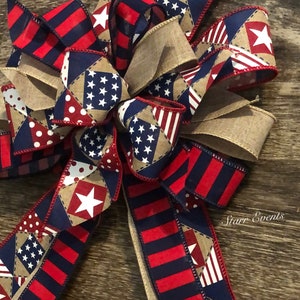 May include: A red, white, and blue patriotic bow with a burlap center. The bow is made of various fabrics with stars and stripes patterns. The bow is tied with a red ribbon and has a "Starr Events" label.
