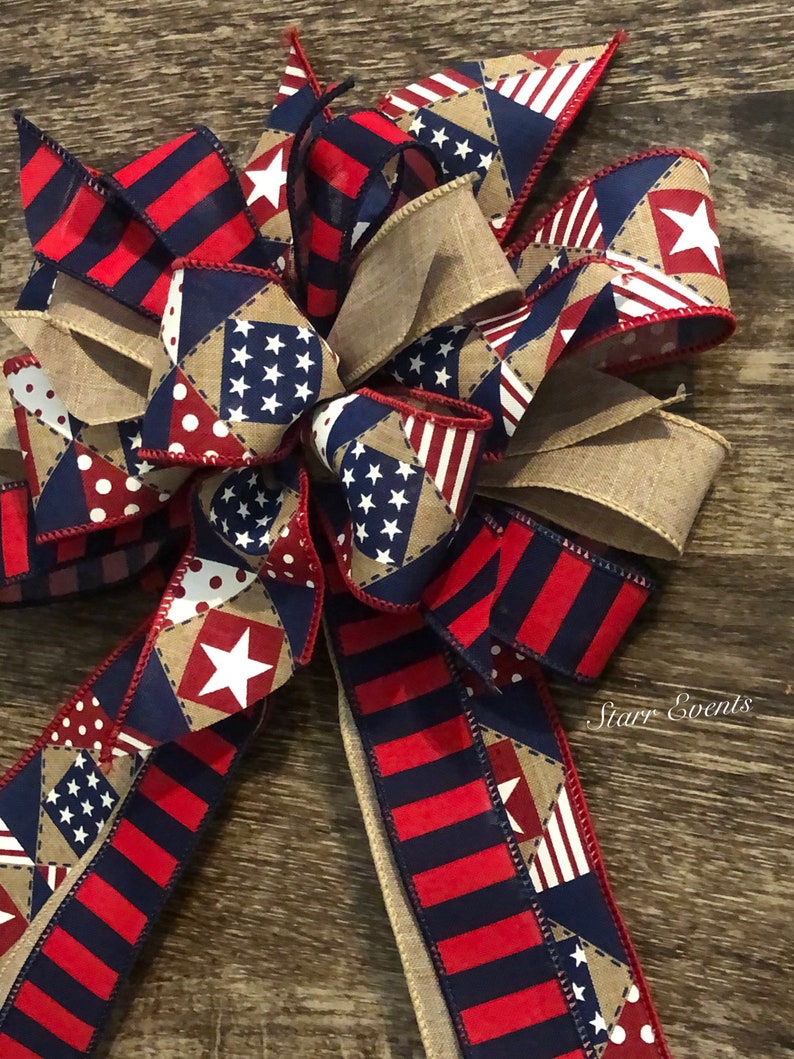 May include: A red, white, and blue patriotic bow with a burlap center. The bow is made of various fabrics with stars and stripes patterns. The bow is tied with a red ribbon and has a "Starr Events" label.