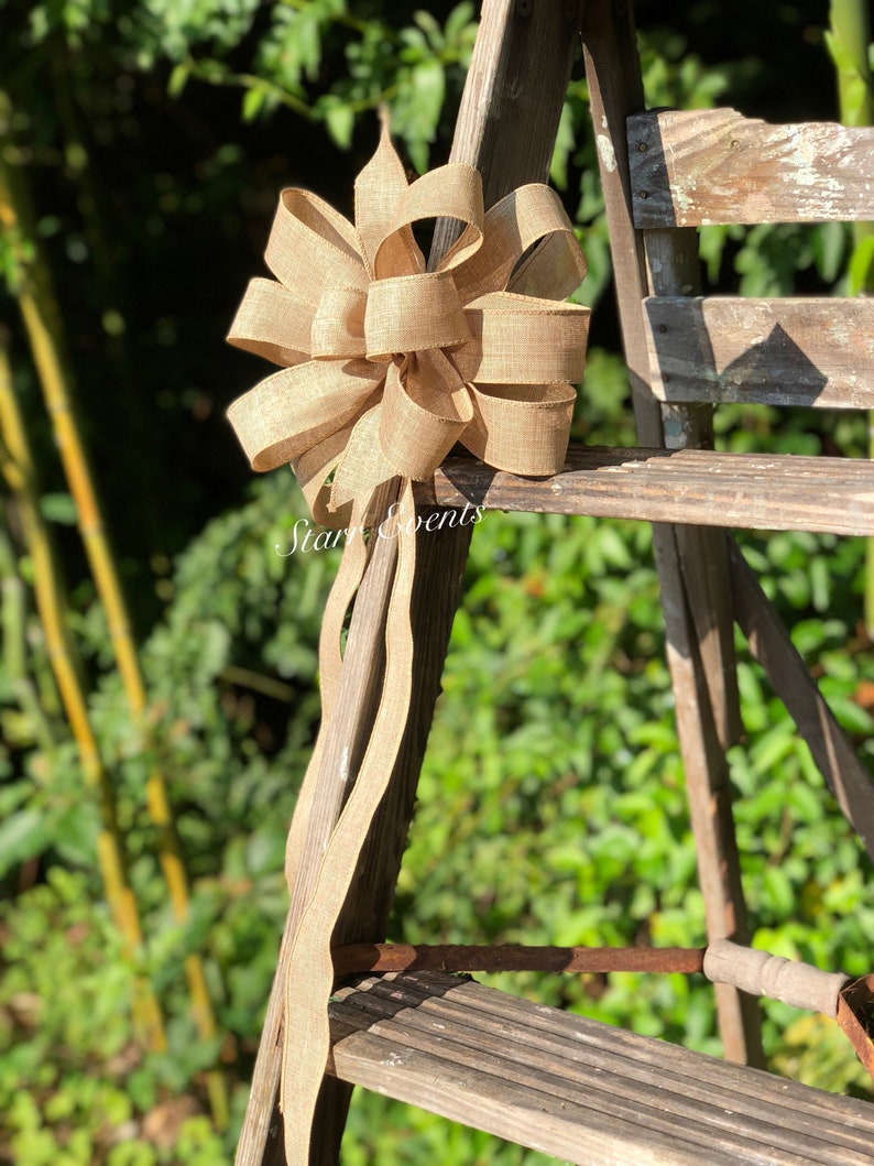 May include: A large, brown burlap bow with a long tail, tied to a wooden ladder. The bow is tied in a classic, symmetrical style with multiple loops.
