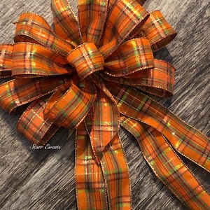 May include: A large orange, green, and gold plaid bow with a shimmery finish. The bow has long tails and is tied with a knot in the center. The bow is on a wood background.