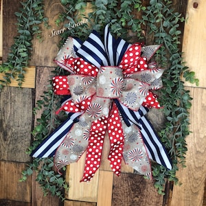May include: A patriotic bow featuring red and white polka dots, navy blue stripes, and a beige ribbon with firework and star patterns. The bow is set against a wooden background with green foliage and the text "Starr Events".