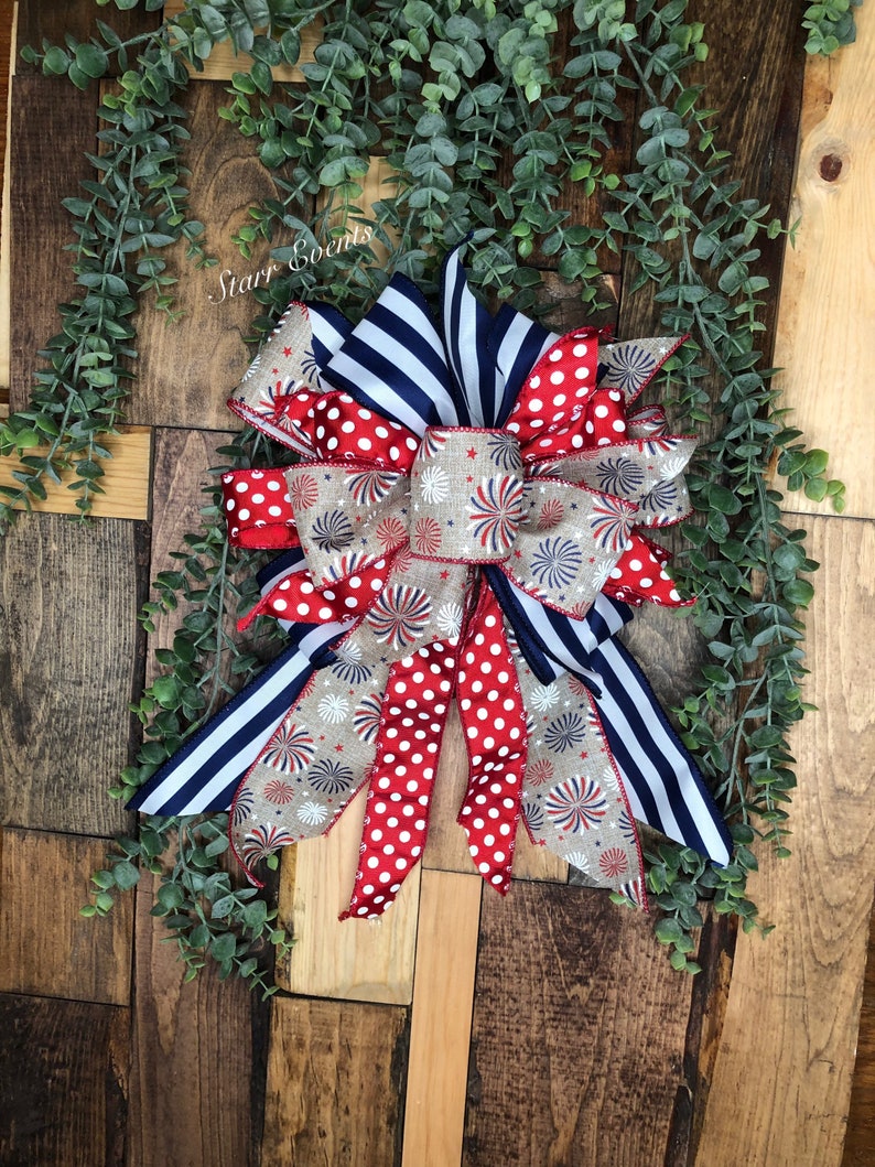 May include: A patriotic bow featuring red and white polka dots, navy blue stripes, and a beige ribbon with firework and star patterns. The bow is set against a wooden background with green foliage and the text "Starr Events".