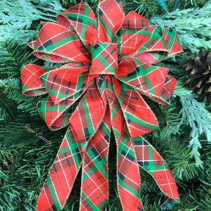 May include: A large, decorative bow with a red, green, and black plaid pattern. The bow is trimmed with gold edging and is set against a backdrop of green pine needles and frosted greenery. A festive holiday decoration.