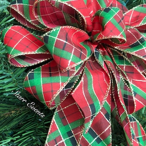 May include: A large, decorative bow with a red, green, and black plaid pattern. The bow is trimmed with gold edging and is set against a backdrop of green pine needles. The bow is likely for holiday or seasonal decor.