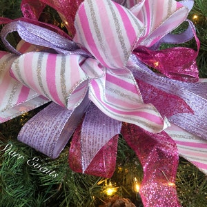 K&ouml;nnte beinhalten: Ein rosa-lila Weihnachtsbaumspitzen-Schleife mit silbernem Glitzer. Die Schleife besteht aus drei verschiedenen Schleifenb&auml;ndern: einem rosa-wei&szlig; gestreiften Band, einem einfarbigen rosa Band mit Glitzer und einem einfarbigen lila Band mit Glitzer.