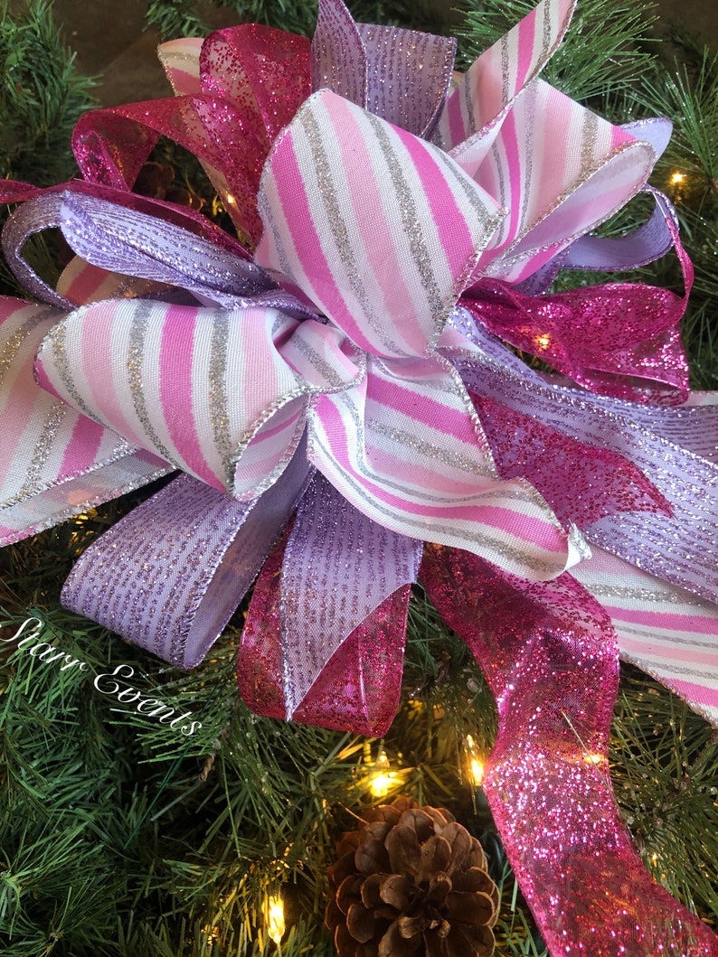 K&ouml;nnte beinhalten: Ein rosa-lila Weihnachtsbaumspitzen-Schleife mit silbernem Glitzer. Die Schleife besteht aus drei verschiedenen Schleifenb&auml;ndern: einem rosa-wei&szlig; gestreiften Band, einem einfarbigen rosa Band mit Glitzer und einem einfarbigen lila Band mit Glitzer.
