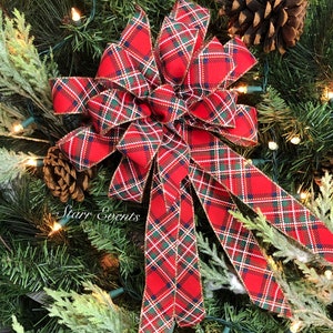 May include: A festive Christmas bow with a red plaid pattern, accented with green and white lines. The bow is set against a backdrop of evergreen branches, pinecones, and twinkling lights, creating a holiday atmosphere. The text "Starr Events" is visible.