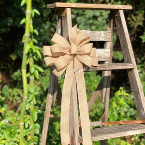 May include: A large, brown burlap bow with two long tails. The bow is tied to a wooden ladder, and the background is a green bush.