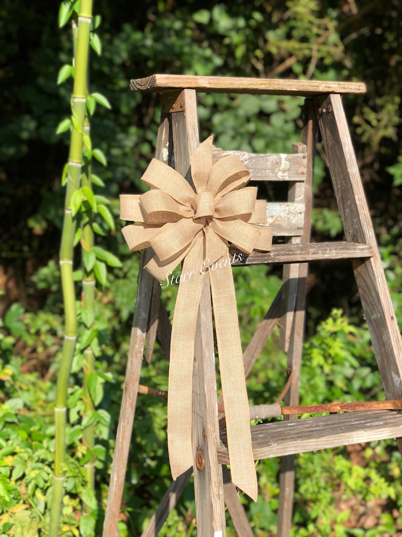 May include: A large, brown burlap bow with two long tails. The bow is tied to a wooden ladder, and the background is a green bush.