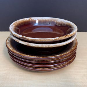 May include: A stack of six brown ceramic bowls with a cream-colored rim. The bowls are stacked on top of each other, with the top two bowls slightly angled. The bowls are on a light brown surface.