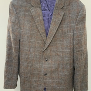 May include: A brown and gray plaid blazer featuring a two-button front closure, notched lapels, and two flap pockets. The interior lining is a vibrant purple fabric with a repeating geometric pattern, visible at the collar and front opening.