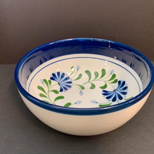 May include: A round ceramic bowl with a cream-colored base and a thick dark blue rim. The interior features a hand-painted floral pattern with blue petals and green leaves, while the exterior remains a solid cream color.