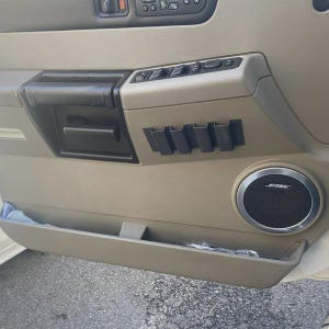 May include: Tan car door panel with a black handle, power window controls, and a Bose speaker. The panel has a storage compartment and small black holders. The door is tan with black accents.