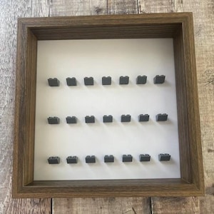 May include: A brown wooden shadow box display frame with a white background and 24 gray plastic slots for displaying Lego bricks.