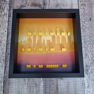 May include: A framed art piece featuring a cityscape silhouette in shades of orange and yellow, with yellow building blocks arranged to mimic skyscrapers. The frame is black, and the background has a gradient effect.