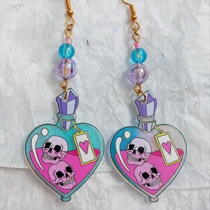May include: A pair of heart-shaped glass bottle earrings with a pink liquid and two skulls inside. The bottles have a purple stopper and a pink tag with a heart on it. The earrings are attached to gold chains with blue and purple beads.