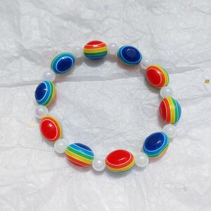May include: A vibrant bracelet with alternating rainbow-striped and solid blue and red beads. The bracelet also includes small, round, white beads. The beads are strung together to form a circular shape.
