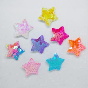 Peut inclure: Une collection de huit embellissements en forme d'étoile de différentes couleurs. Chaque étoile est recouverte de paillettes, créant un effet scintillant. Les couleurs incluent le jaune, le rose, le violet, l'orange et le bleu. Idéal pour les loisirs créatifs.