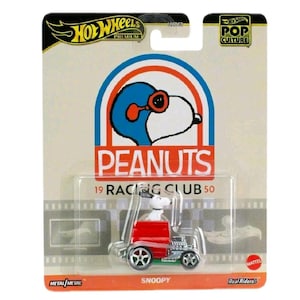 Op de afbeelding: Een Hot Wheels Premium gegoten auto met Snoopy in een rood voertuig. De verpakking bevat het Peanuts-logo, de tekst "19 Racing Club 50" en de Hot Wheels- en Pop Culture-logo's. De auto heeft zilveren wielen en een groene basis.