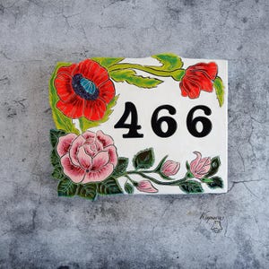 Puede incluir: Una placa de cerámica rectangular para número de casa con los dígitos negros 466 sobre un fondo blanco. La placa está decorada con amapolas rojas y rosas rosadas pintadas a mano con hojas verdes en los bordes superior e inferior.