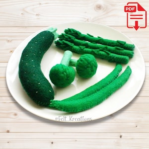 May include: A set of felt food items, including a courgette, asparagus, broccoli, and green beans, arranged on a white plate. The felt food items are green and are designed to look like real vegetables.