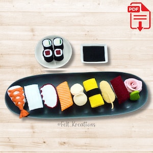 Puede incluir: Un conjunto de sushi de fieltro con varios tipos de sushi, incluyendo salmón, atún y camarones. El conjunto también incluye un pequeño plato de salsa de soja y una rosa de fieltro rosa.