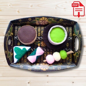 May include: A black tray with a patterned design holds a variety of felt food items, including a green tea cup, a brown cake, a green and white mochi, a pink and white mochi, and a skewer of three green and white mochi.