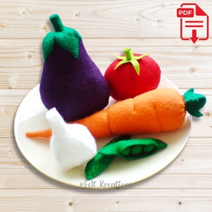 Puede incluir: Un plato blanco con una berenjena morada, un tomate rojo, una zanahoria naranja, un ajo blanco y una vaina de guisante verde. Las verduras están hechas de fieltro y están dispuestas en un plato blanco. La imagen está etiquetada "@Felt_Kreations".