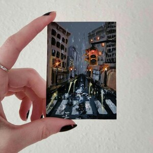 May include: Miniature painting of a city street scene in the rain. The artwork depicts a crosswalk, buildings, and traffic lights. The colour palette includes shades of grey, brown, and yellow, creating a moody atmosphere. The painting is held by a hand.