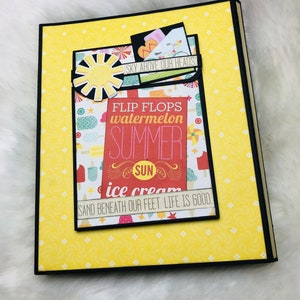 May include: A yellow scrapbook with a black border and a pocket on the front. The pocket has a colourful design with the text "FLIP FLOPS watermelon SUMMER SUN ice cream SAND BENEATH OUR FEET. LIFE IS GOOD."