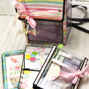 May include: A set of colorful scrapbook pages with a pink ribbon and a clear plastic cover. The pages feature various designs, including dinosaurs, balloons, and a vintage suitcase. The pages are labeled with the words "You are Dino-mite", "Party Time", and "Memory Time".